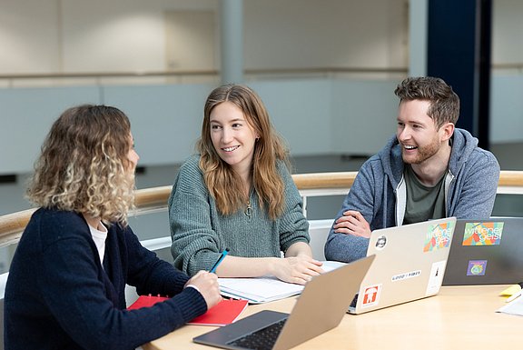 Drei Studierende sitzen mit Laptops am Tisch und bilden eine Lerngruppe