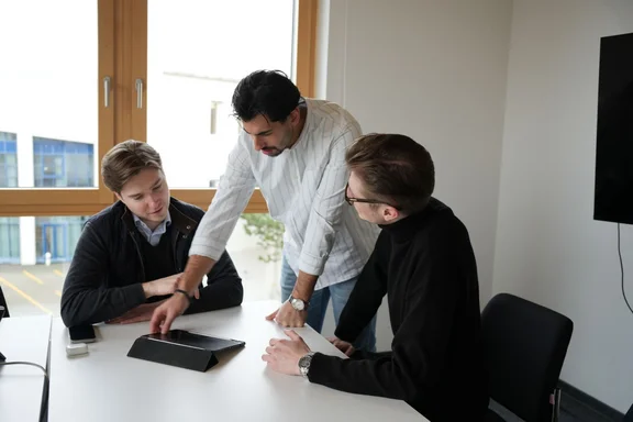 Drei Studierende sitzen zusammen an einem Konferenztisch und schauen gemeinsam auf ein Tablet.
