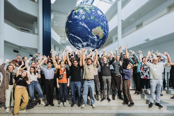 Zu sehen ist ein Gruppenfoto in der Eingangshalle des Hauptgebäudes, in dem alle die Arme in die Luft werfen, über ihren Köpfen das große Modell einer Weltkugel