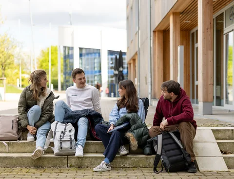 Aktuelles vom Campus (Foto: UW/H | Michael Schwettmann) Eine Gruppe Studierender sitzt auf den Campustreppen neben dem Holzgebäude der Uni Witten/Herdecke. Sie sind in ein Gespräch vertieft.