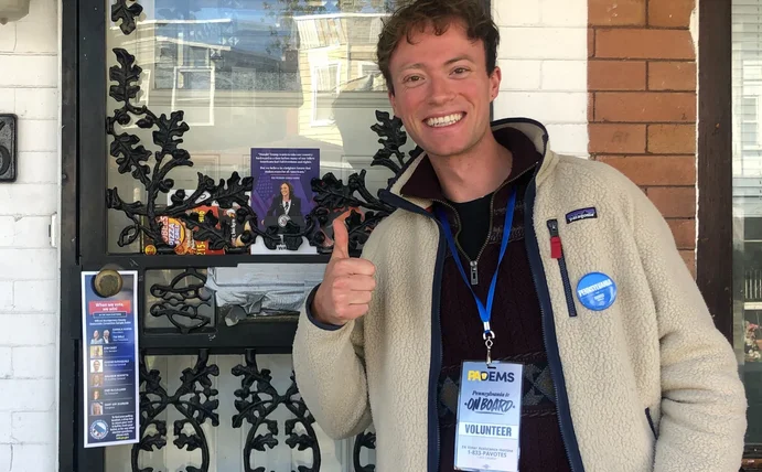 Eine Person zeigt den Daumen hoch vor einer Haustür mit Wahlkampfmaterial.