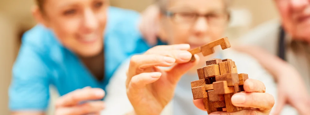 Eine ältere Frau versucht ein Holzpuzzel zu lösen. Neben ihr im Hintergrund sind zwei weitere Personen, die ihr zusehen.