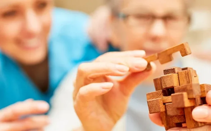 Eine ältere Frau versucht ein Holzpuzzel zu lösen. Neben ihr im Hintergrund sind zwei weitere Personen, die ihr zusehen.