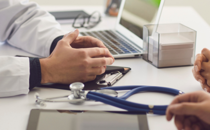 Ein Arzt und ein Patient sitzen sich am Tisch gegenüber. Der Arzt trägt einen weißen Kittel. Zwischen ihnen liegt ein Stethoskop.