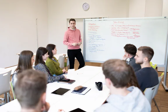 Die Wissenschaftler:innen der UW/H arbeiten in interdisziplinären Forschungsprojekten fakultätsübergreifenden zusammen (Foto: UW/H | Michael Schwettmann) Ein junger Mann steht an einem Whiteboard und schaut in die Gesichter junger Menschen, die im Kreis an einem Tisch vor ihm sitzen.