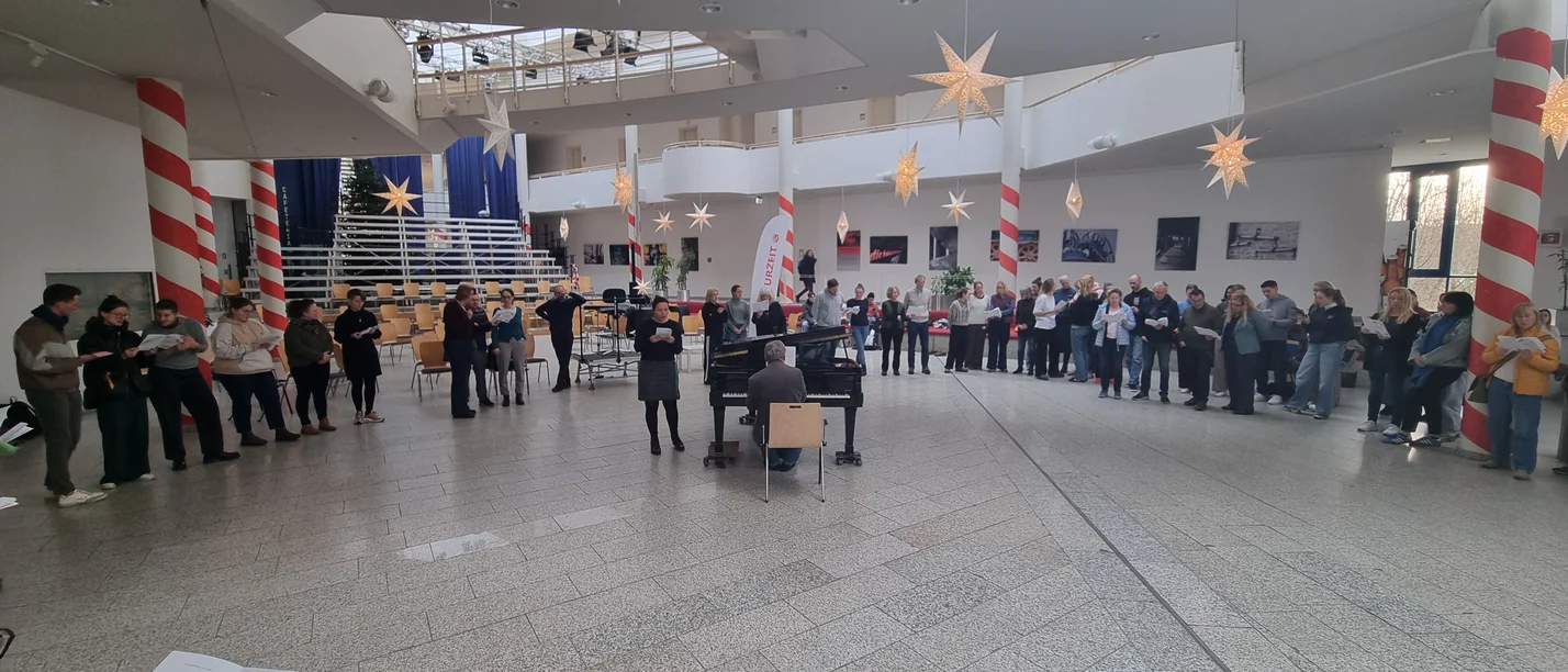 A group of people singing together in the UW/H hall, which is decorated for Christmas. A man is sitting at the piano, accompanying them