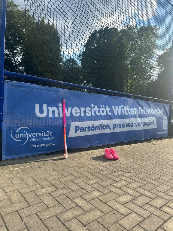 Foto eines blauen Werbebanners am Spielfeldrand. Davor stehen pinke Sportschuhe und ein Hockeyschläger