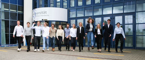 Eine Gruppe Studierender läuft vom Gebäude-Eingang der Universität lachend auf die Kamera zu.