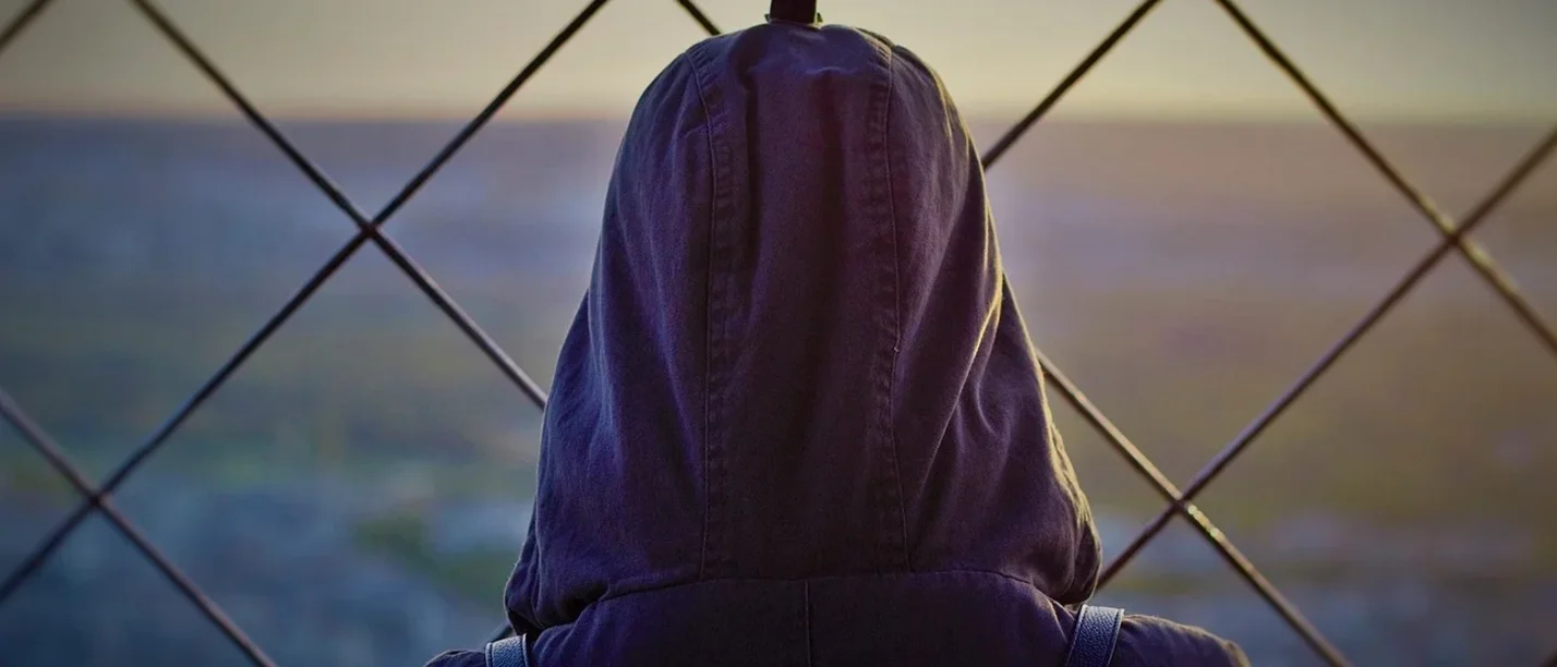 Ein Mensch steht mit dem Rücken zur Kamera und schaut durch die Löcher eines Zauns in die Ferne.