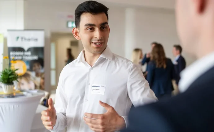 Ein junger Mann unterhält sich mit einem anderen Mann. Im Hintergrund ist sind ein Stehtisch und weitere Menschen.