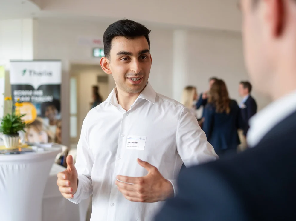 Ein junger Mann unterhält sich mit einem anderen Mann. Im Hintergrund ist sind ein Stehtisch und weitere Menschen.