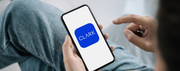 Mit der Smartphone-App CLARK können Kund:innen ihre Versicherungen digital verwalten. (Foto: Canva; Grafik: CLARK) Eine sitzende Person hält ein Smartphone in den Händen. Der Fokus liegt auf dem Smartphone.