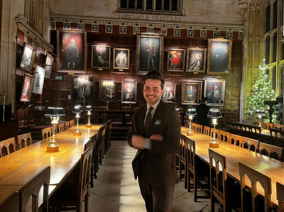 Dass Sami einmal einen akademischen Weg einschlagen würde, war für ihn lange Zeit undenkbar. Als er 2024 das erste Mal für ein Praktikum nach Oxford kam, konnte er sein Glück kaum fassen. (Foto: privat) Sami Barkal steht in einem historischem Saal der Oxford University. Hinter ihm hängen Porträts an der Wand. In der hinteren rechten Ecke steht ein beleuchteter Weihnachtsbaum.