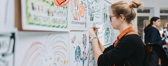 WittenLab der UW/H: Forschung an der Schnittstelle von Philosophie, Kommunikations-, Geschichts-, Kunst- und Musikwissenschaften, Soziologie und Psychologie (Foto: UW/H | Michael Schwettmann) Eine Frau schreibt etwas auf Plakate, die an Metaplanwänden hängen.