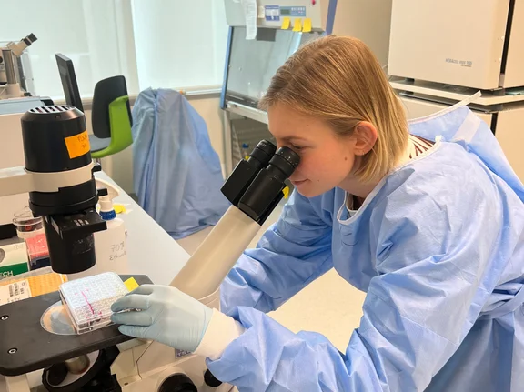 A female student, wearing a blue lab coat and gloves, is working intently at a microscope in a research laboratory.