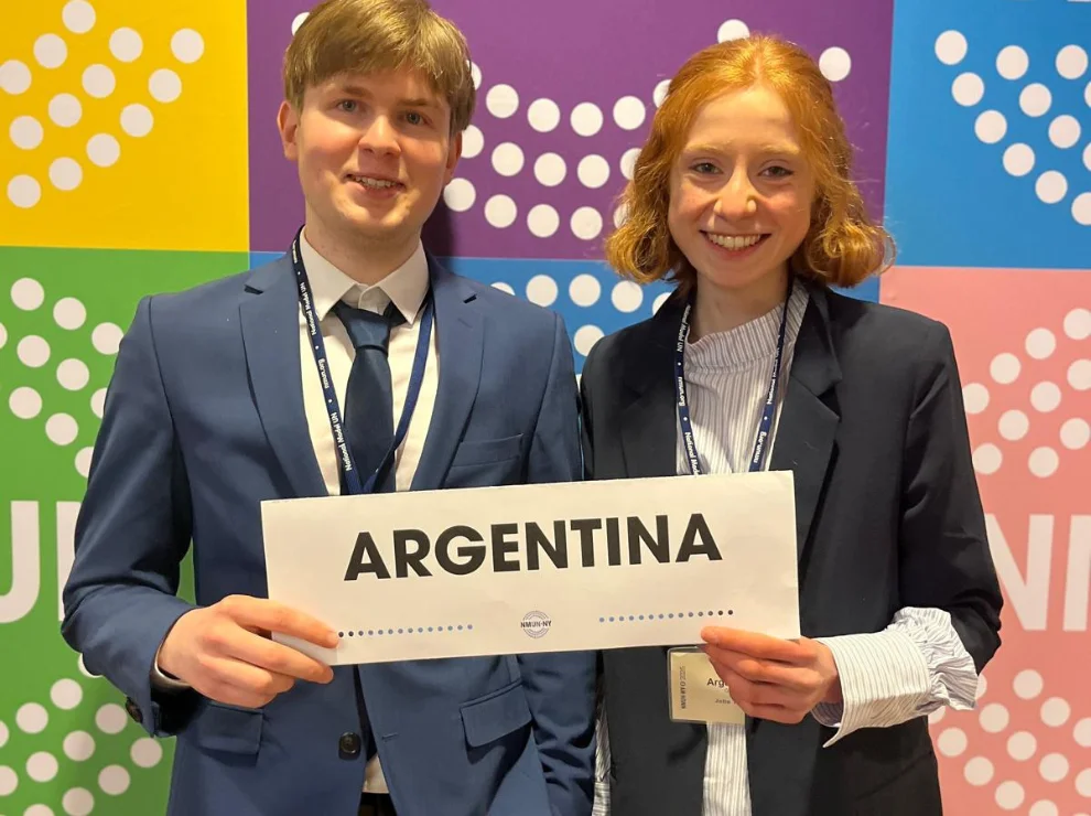 Zwei Studierende stehen vor einer bunten Wand und halten ein Schild mit der Aufschrift "Argentina" hoch.