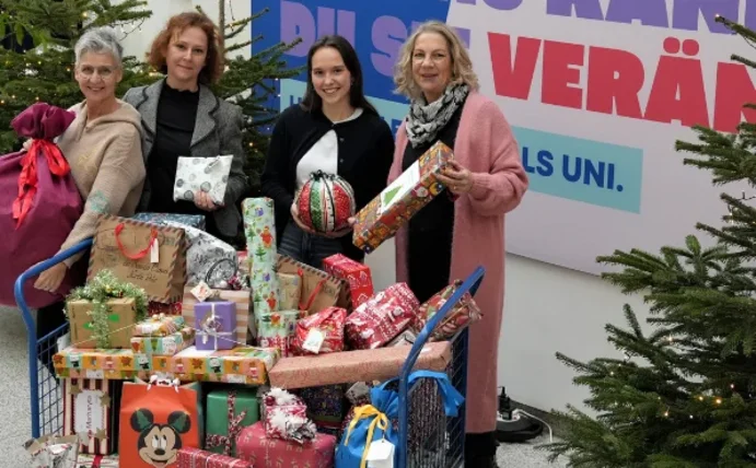 Vier Frauen stehen zwischen Tannenbäumen und halten Weihnachtsgeschenke in den Händen. Vor ihnen steht ein Transportwagen mit weiteren Päckchen.