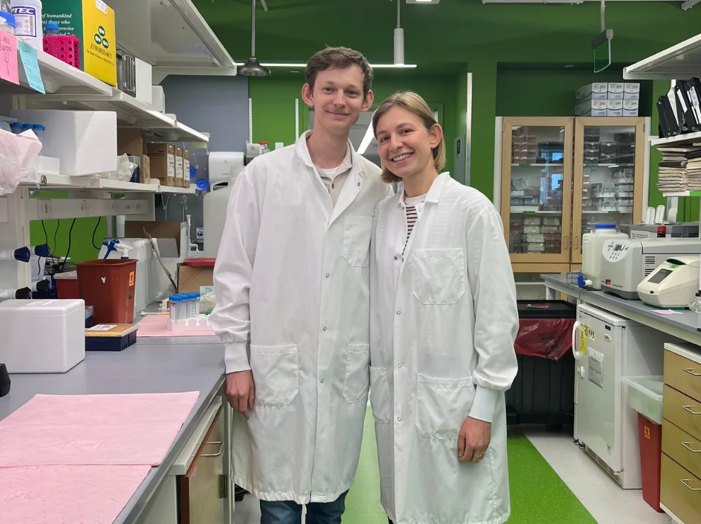 Two medical students in white lab coats are standing side by side in a modern research laboratory, smiling at the camera.