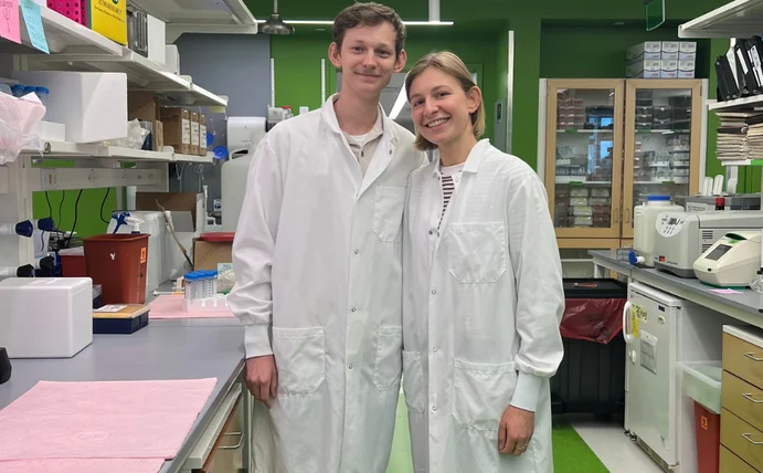 Two medical students in white lab coats are standing side by side in a modern research laboratory, smiling at the camera.