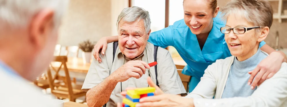 Eine Pflegerin hat den Arm um die Schultern eines Mannes und einer Frau gelegt. Alle lachen, der Mann hält einen Bauklotz in der Hand, die Frau rpckt einen Turm aus Bauklötzen zurecht.