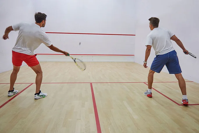 Beim Squash-Spiel auspowern (Foto: Canva) Zwei Männer spielen in einer Sporthalle Squash. Einer von ihnen schlägt gerade den Ball gegen die Wand.