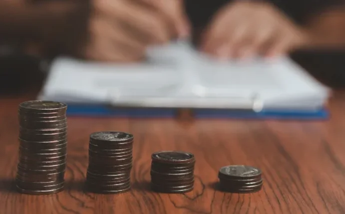 Vorne sieht man aufgestapelte Münzen, im Hintergrund Hände mit einem Stift in der Hand. 