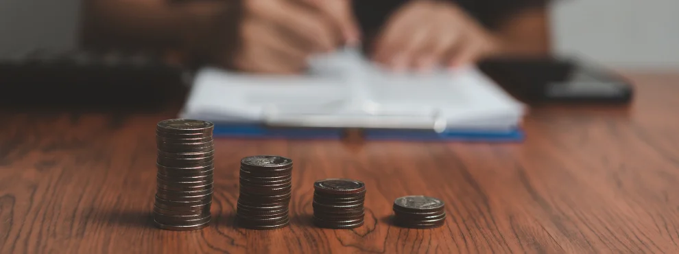 Vorne sieht man aufgestapelte Münzen, im Hintergrund Hände mit einem Stift in der Hand. 