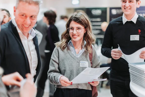 Wertvolle Verbindungen (Foto: UW/H | Michael Schwettmann) Tagungssituation: Zwei Männer und eine Frau melden sich für ein Event an. Sie halten Unterlagen in den Händen und lächeln.