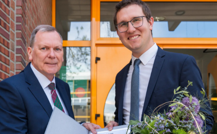 Zwei Männer lächeln in die Kamera und halten gemeinsam eine Urkunde hoch. Der Mann rechts hat außerdem einen Blumenstrauß in der Hand.