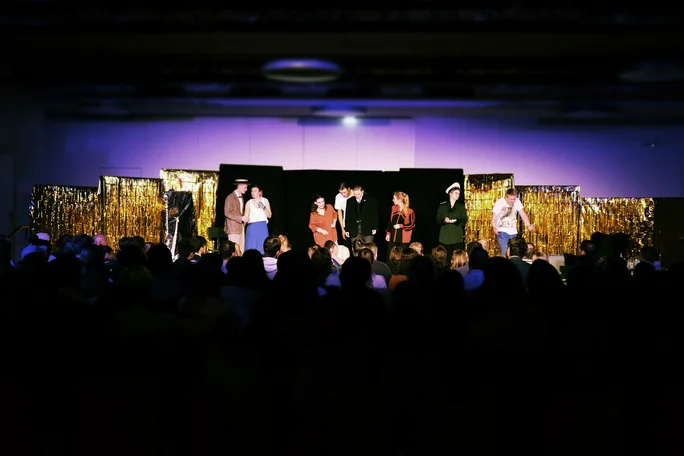 Theatre performance at the UW/H (Photo: UW/H). It is very dark in the theatre hall, only the stage is illuminated. The silhouettes of the audience can be seen in the foreground, while acting students in their costumes stand on the stage in the background.