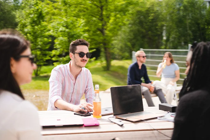 Mitten im Grünen: Lernen am UW/H-Campus (Foto: UW/H | Johannes Buldmann) Eine Gruppe Studierender sitzt gemeinsam an einem Tisch auf dem Campus. Da es sonnig und warm ist, tragen sie Sonnenbrillen und sommerliche Kleidung. Zwischen ihnen stehen ein Laptop, Lernunterlagen und Getränke.