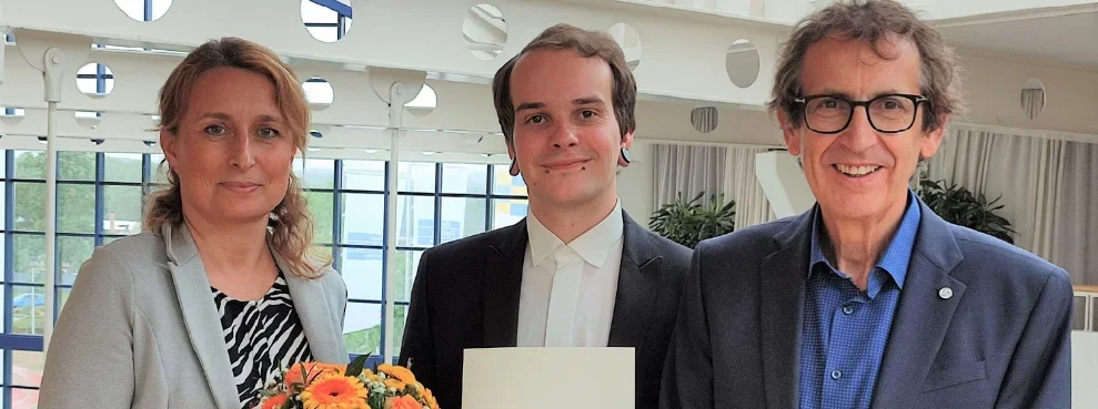 Drei Personen lächeln in die Kamera. Die Person in der Mitte hält eine Urkunde in den Händen. Die Person links überreicht der Person in der Mitte einen Blumenstrauß.