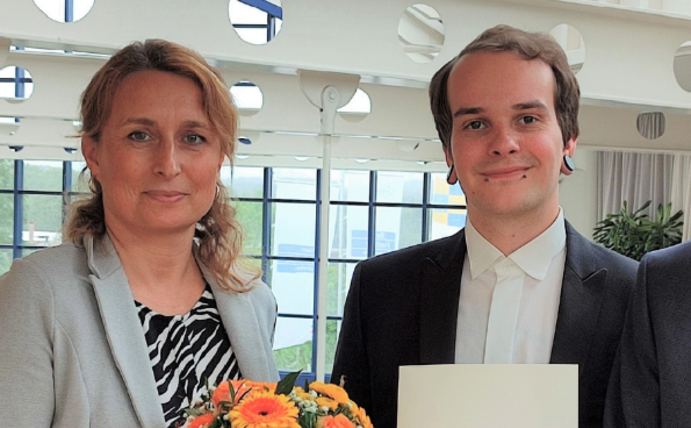 Drei Personen lächeln in die Kamera. Die Person in der Mitte hält eine Urkunde in den Händen. Die Person links überreicht der Person in der Mitte einen Blumenstrauß.