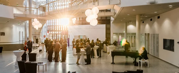 In einer lichtdurchfluteteten Halle stehen Menschen an Stehtischen und unterhalten sich.
