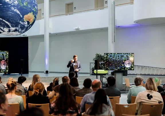An der UW/H erforschen wir, wie wir neue Narrative für planetare Bildung schaffen können. (Foto: UW/H | Michael Schwettmann) Ein Mann steht auf einer Bühne und spricht vor Publikum, hinter ihm ragt eine große Weltkugel ins Bild.