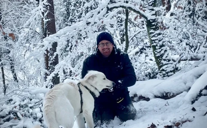 A man kneels in a winter landscape with trees, in front of him stands a white dog.