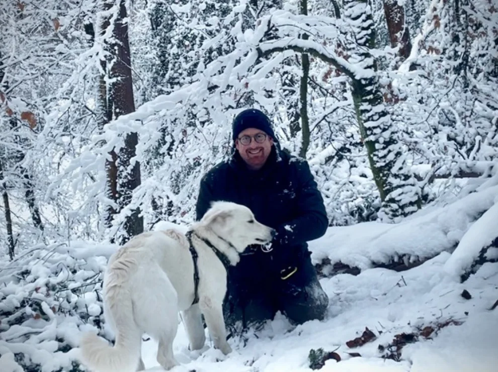 Ein Mann kniet in einer Winterlandschaft mit Bäumen, vor ihm steht ein weißer Hund.