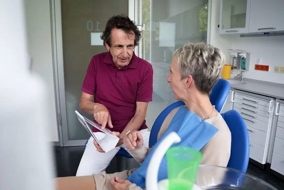 In den Ambulanzen der UW/H arbeiten arbeiten Therapeut:innen, Ärzt:innen und zahnmedizinische Fachangstellte eng mit weiteren Fachleuten zusammen. (Foto: UW/H) Ein Zahnarzt zeigt einer Patientin etwas auf einem Blatt Papier.