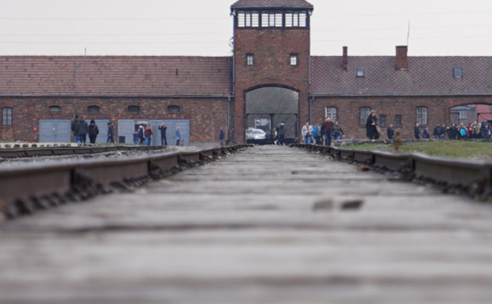 Im Vordergrund sieht man verschwommene Bahnschienen. Im Hintergrund das KZ Auschwitz, drumherum ganz klein ein paar Menschen.