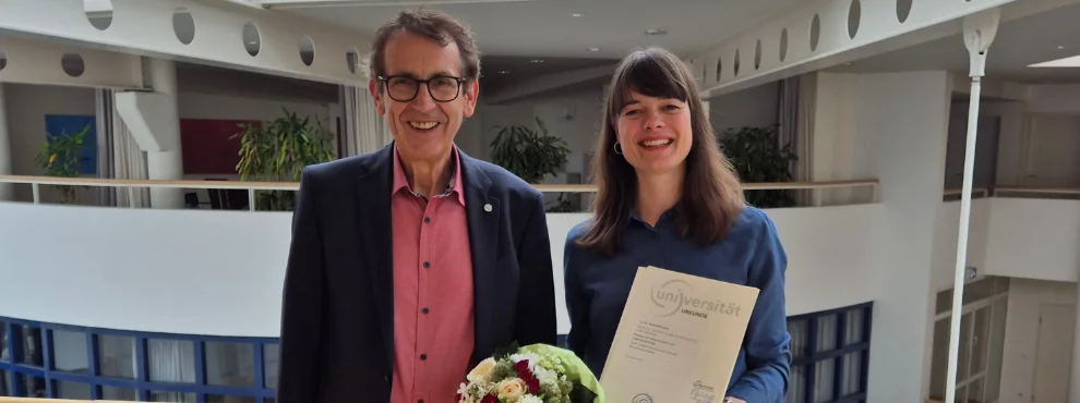Ein Mann und eine Frau stehen auf vor einer Brüstung der Universität Witten/Herdecke und lächeln in die Kamera. Die Frau hält ihre Berufungsurkunde in der Hand. 
