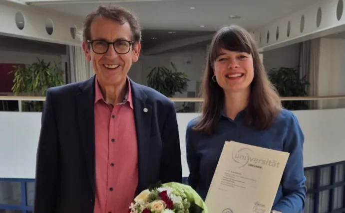 Ein Mann und eine Frau stehen auf vor einer Brüstung der Universität Witten/Herdecke und lächeln in die Kamera. Die Frau hält ihre Berufungsurkunde in der Hand. 