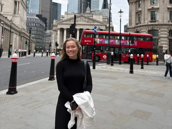 Eine junge Frau steht auf einem großen Platz in London. Hinter ihr fährt ein typischer roter Doppeldecker-Bus vorbei.