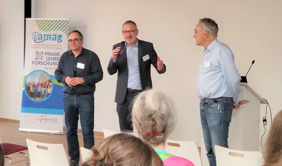 Drei Männer stehen vor einem Publikum, der Mann in der Mitte spricht in ein Mikrofon.