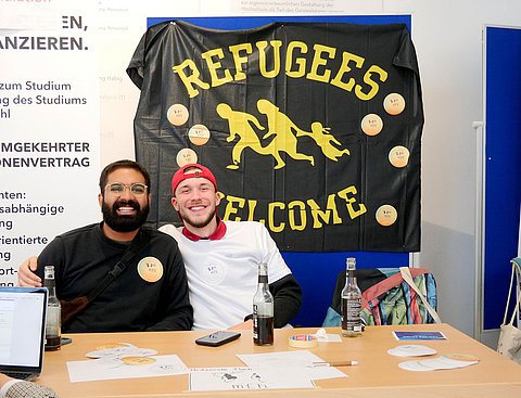 Zwei Studierende sitzen an einem Tisch, halten sich in den Armen und lächeln in die Kamera. Hinter ihnen hängt ein Banner mit der Aufschrift "Refugees welcome". Die Studierenden informieren an ihrem Stand über ihr Engagement.