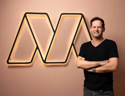 Student companies: UW/H graduates found further education platform Masterplan.com. (Photo: Founder Stefan Peukert | Anke Peters) A man stands smiling and with his arms folded in front of an apricot-coloured man. Next to him on the wall is the logo ("M") of his company.
