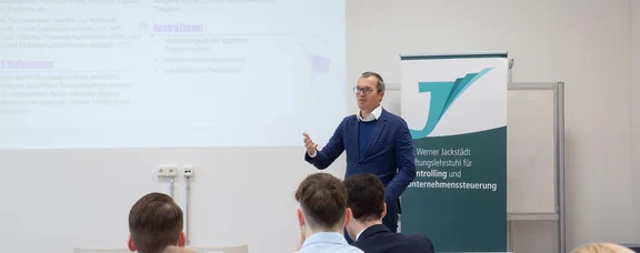 We bring practical experience into the seminar room (Photo: UW/H | Miriam Kreimeyer) A man in a suit is standing in a lecture theatre, speaking to students. A presentation is being shown on the wall behind him.