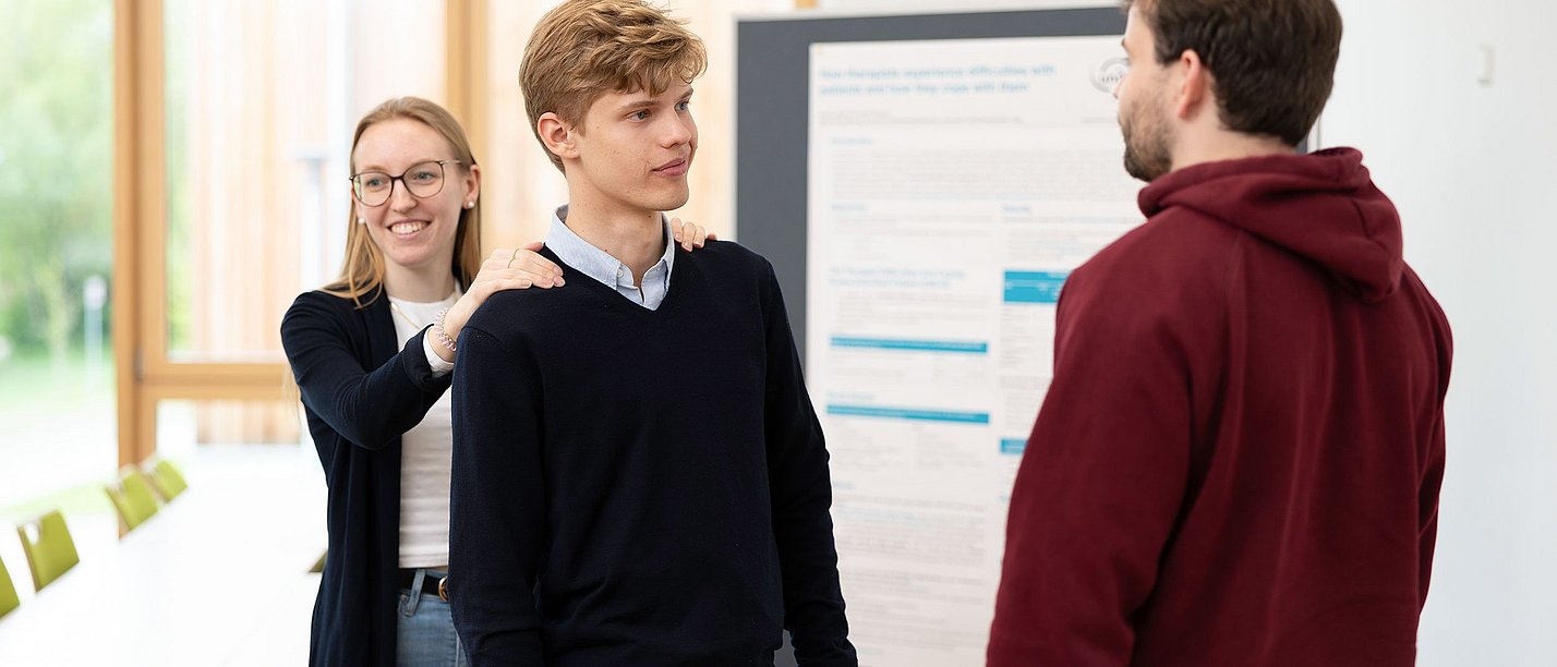 In der Online-Infoveranstaltung können Studieninterssierte den Bachelor-Studiengang Psychologie entdecken. (Foto: UW/H | Michael Schwettmann) Ein Student steht mit dem Rücken zu einer Studentin und schaut einen anderen Studenten an. Die Studentin hat ihm die Hände auf die Schultern gelegt.