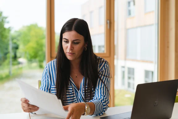 A student is sitting in front of her laptop, holding a sheet of paper in her hands.