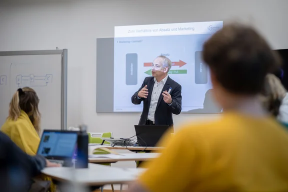 Course (Photo: UW/H | Michael Schwettmann) A lecturer is standing in front of students in a seminar room. A presentation is being shown behind him.