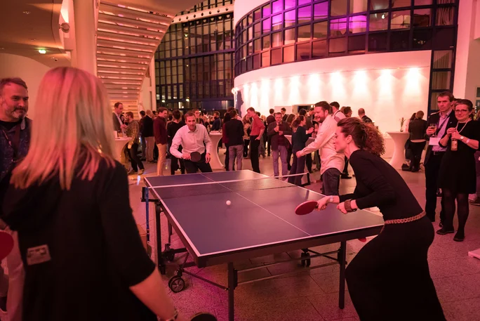Celebrating together at the alumni gathering (Photo: UW/H | Florian Danner) Two people are playing table tennis in an event foyer. In the background, there are other people watching or chatting.
